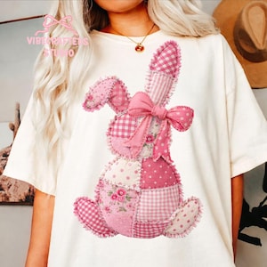 Könnte beinhalten: Cremefarbenes T-Shirt mit einem Patchwork-Hasen-Design in verschiedenen Rosatönen. Der Hase hat eine große Schleife und besteht aus verschiedenen gemusterten Stoffen, darunter Vichy, Tupfen und Blumendrucke. Der Text "Vibe Crafters Studio" ist sichtbar.