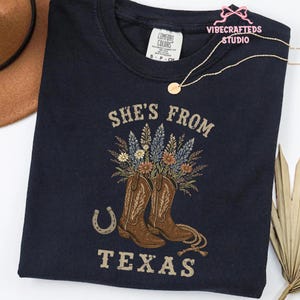 May include: Navy blue t-shirt with the text "She's From Texas". The design features brown cowboy boots filled with flowers, a horseshoe, and rope detailing. The shirt has a Comfort Colors tag.