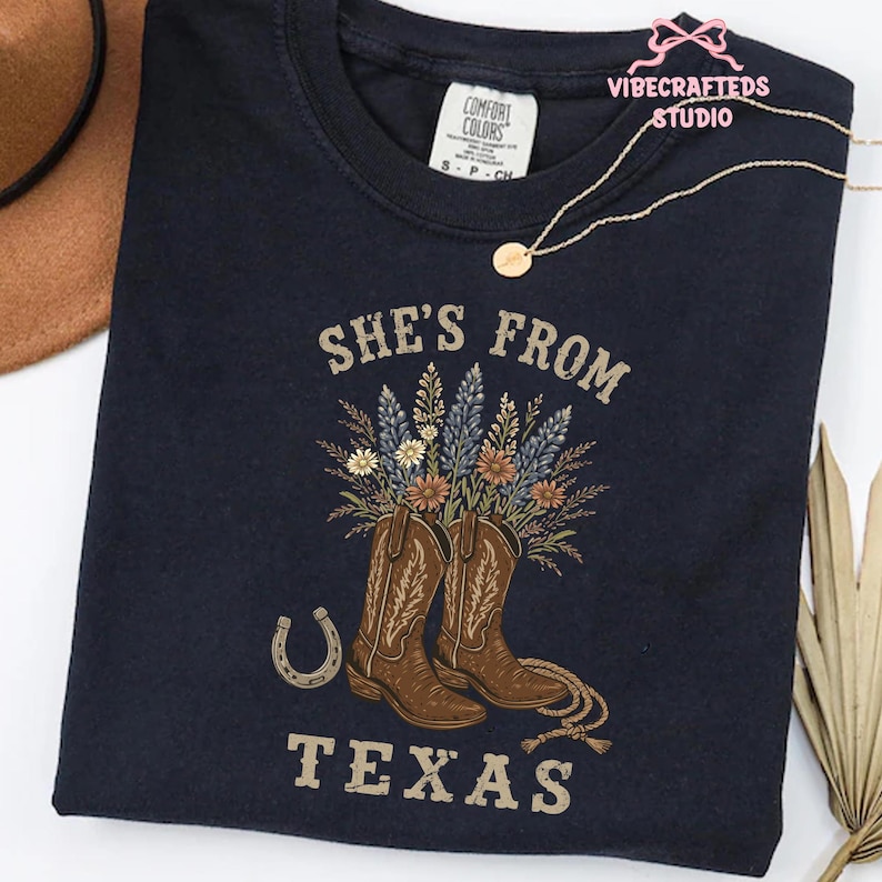 May include: Navy blue t-shirt with the text "She's From Texas". The design features brown cowboy boots filled with flowers, a horseshoe, and rope detailing. The shirt has a Comfort Colors tag.