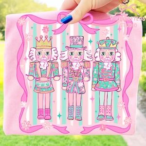 Pode incluir: Uma camiseta rosa com um desenho de três figuras de quebra-nozes. Os quebra-nozes usam roupas rosa, verde-azulado e douradas com estampas florais. O fundo tem listras rosa e verde-azulado, com uma fita rosa e o texto "Vibe Crafted Studio".