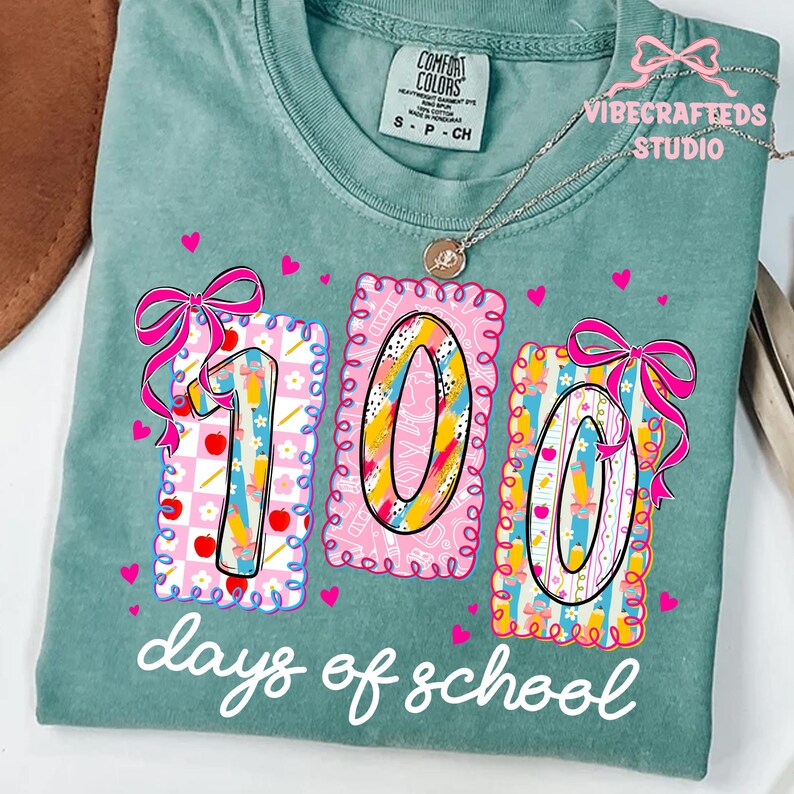 May include: A teal Comfort Colors t-shirt with a colorful graphic celebrating 100 days of school. The design features the numbers "100" in a decorative frame with bows, hearts, and school-themed elements. The text "days of school" is written in a script font.