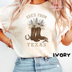 May include: Ivory-colored t-shirt featuring a graphic with brown cowboy boots, a horseshoe, and floral elements. The text "SHE'S FROM TEXAS" is displayed above the boots. The shirt has a relaxed fit.