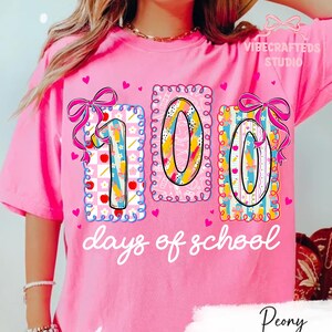 May include: Pink t-shirt with a colorful "100 days of school" graphic. The design features the numbers 1, 0, and 0 in decorative frames with bows and hearts. The text "days of school" is written in a cursive font.