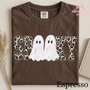 May include: A dark brown t-shirt featuring the word SPOOKY in a collegiate font. The letters S, P, K, and Y have a tan and black leopard print pattern. The two O letters are replaced by white lace ghosts with black eyes. The shirt has a crew neckline.