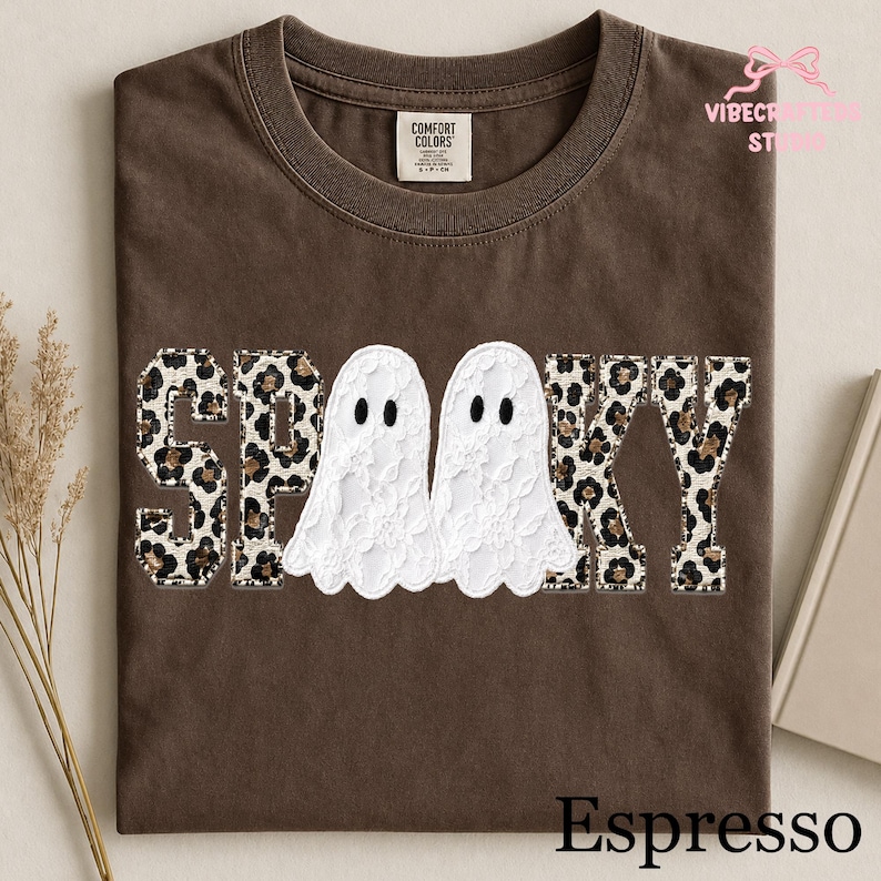May include: A dark brown t-shirt featuring the word SPOOKY in a collegiate font. The letters S, P, K, and Y have a tan and black leopard print pattern. The two O letters are replaced by white lace ghosts with black eyes. The shirt has a crew neckline.