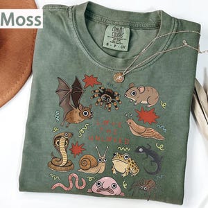 May include: A moss green t-shirt with a design of various illustrated creatures, such as a bat, spider, mouse, and snake. The text "Love the Unloved" is printed on the shirt. A silver necklace with a pendant is draped across the shirt.