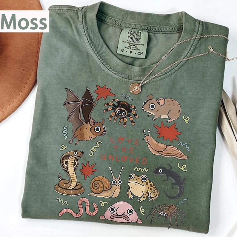 May include: A moss green t-shirt with a design of various illustrated creatures, such as a bat, spider, mouse, and snake. The text "Love the Unloved" is printed on the shirt. A silver necklace with a pendant is draped across the shirt.