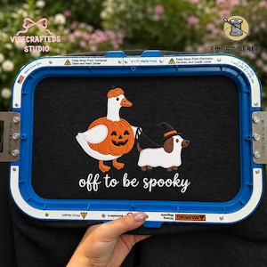 Op de afbeelding: Een zwart stuk stof in een blauwe borduurring met een geborduurde witte gans in een oranje pompoenkostuum en een bruin-witte hond met een zwarte heksenhoed, met daaronder de witte tekst "off to be spooky".