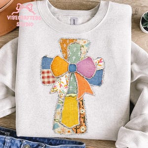 May include: A light gray sweatshirt featuring a colorful, patchwork cross design. The cross is adorned with a large bow and various fabric patterns, including plaid and floral prints. The text "VIBECRAFTED STUDIO" is visible in the upper left corner.