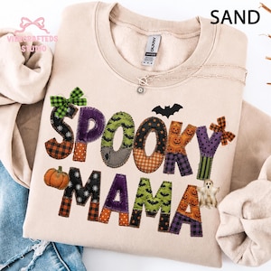 May include: A sand-colored crewneck sweatshirt featuring the text SPOOKY MAMA in a patchwork-style font. The letters are decorated with patterns of pumpkins, bats, spiders, and plaid, accented by small green and orange bows and a ghost graphic.