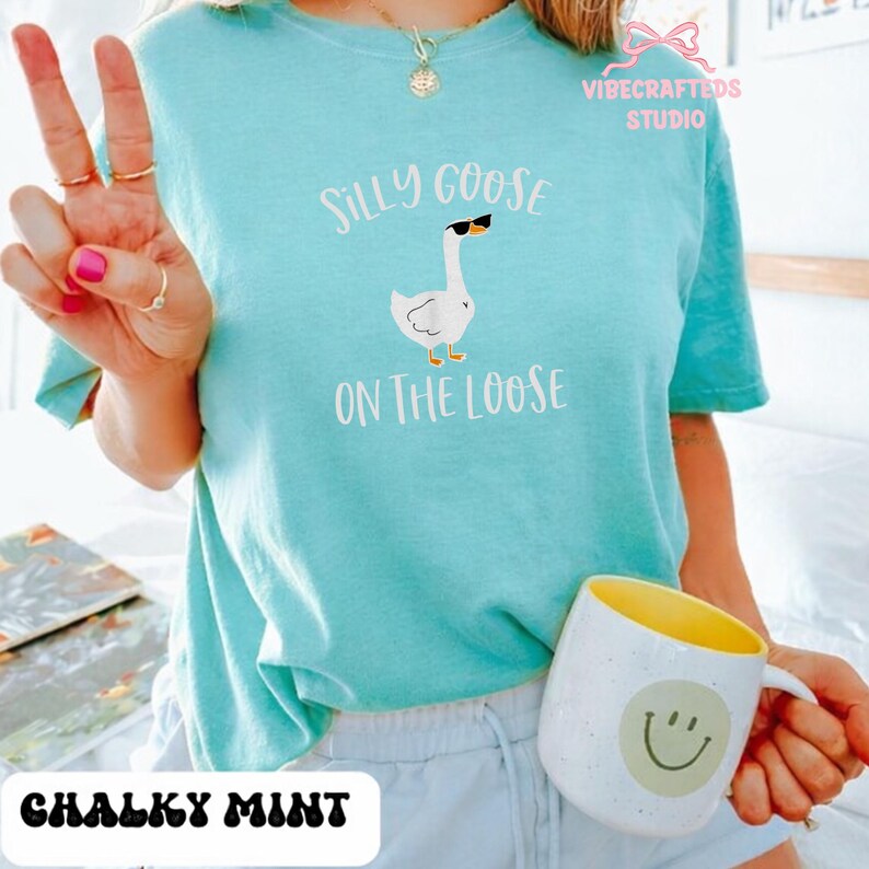May include: A chalky mint-colored t-shirt with the text "Silly Goose On The Loose" and a graphic of a goose wearing sunglasses. A white mug with a yellow interior and a smiley face is also visible.