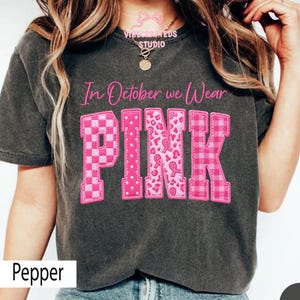 May include: A charcoal gray t-shirt featuring the pink text In October we Wear PINK. The word PINK is displayed in large block letters with varied patterns including checkers, polka dots, leopard print with ribbons, and a plaid design.
