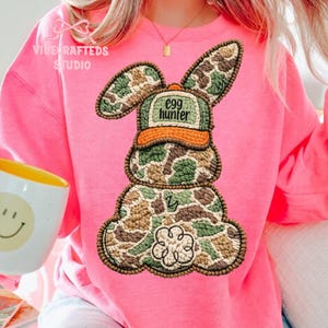 May include: A pink sweatshirt featuring a camouflage bunny design with the text "egg hunter" on the hat. The bunny has a green, brown, and tan pattern. The sweatshirt has the text "Vibe Crafted Studio" in the upper left corner.