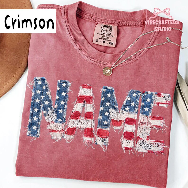May include: Crimson t-shirt with the word "NAME" in a distressed American flag design. The letters feature blue denim with white stars and red and white striped fabric. The shirt has a Comfort Colors tag and a silver necklace.