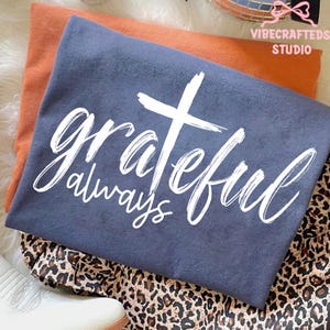 May include: A folded navy blue t-shirt with the words "grateful always" in white script and a white cross graphic. An orange shirt is underneath, and the shirts are on a leopard print fabric.