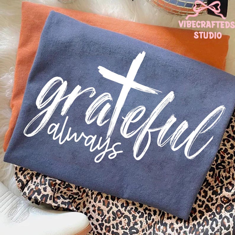 May include: A folded navy blue t-shirt with the words "grateful always" in white script and a white cross graphic. An orange shirt is underneath, and the shirts are on a leopard print fabric.