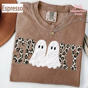 May include: A brown crew neck t-shirt featuring the word SPOOKY in block letters with a leopard print pattern. The two O's are replaced by white lace ghost figures with black oval eyes. The shirt has a Comfort Colors label at the inner neckline.