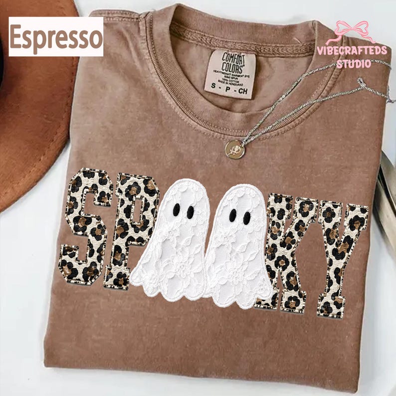 May include: A brown crew neck t-shirt featuring the word SPOOKY in block letters with a leopard print pattern. The two O's are replaced by white lace ghost figures with black oval eyes. The shirt has a Comfort Colors label at the inner neckline.
