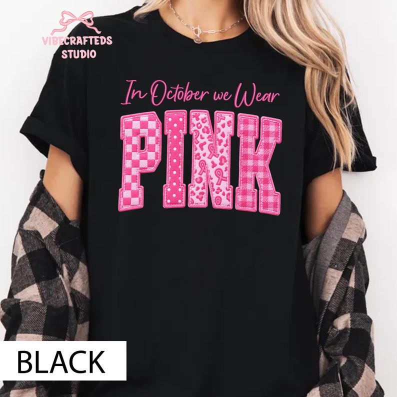 May include: A black short-sleeved t-shirt featuring the pink text In October we Wear PINK. The letters are filled with various patterns including checkers, polka dots, leopard print with ribbons, and plaid. A logo is visible in the top left corner.