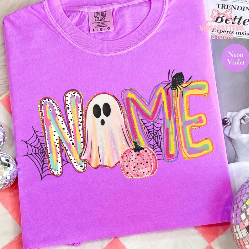 May include: A neon violet t-shirt featuring a graphic print with the word NAME. The letter A is replaced by a ghost, and the letter E is accompanied by a pink pumpkin and a black spider. The letters have a colorful, brushstroke-style pattern.