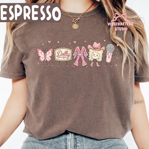 May include: A dark brown short-sleeve t-shirt featuring a graphic print of a pink butterfly, a marquee sign with the text Dolly, a pink jumpsuit, a blonde character in a pink cowboy hat, and a microphone, all holding hands with small stars and hearts.