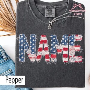 May include: Dark grey t-shirt featuring the word "NAME" in a design resembling the American flag. The letters are blue with white stars and red and white stripes. The shirt has a crew neck and a small "COMFORT COLORS" tag.