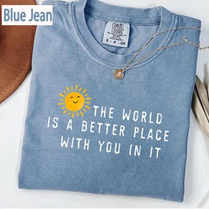 May include: A blue jean colored t-shirt featuring a graphic of a yellow smiling sun and the white text "THE WORLD IS A BETTER PLACE WITH YOU IN IT". The shirt has a crew neckline and a Comfort Colors brand label visible at the inner collar area.