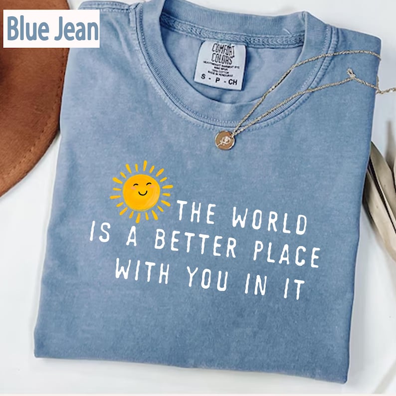 May include: A blue jean colored t-shirt featuring a graphic of a yellow smiling sun and the white text "THE WORLD IS A BETTER PLACE WITH YOU IN IT". The shirt has a crew neckline and a Comfort Colors brand label visible at the inner collar area.
