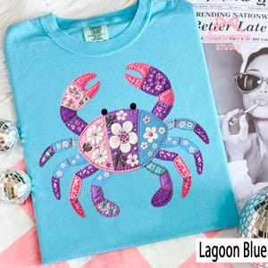 Puede incluir: Una camiseta azul claro con un aplique de cangrejo colorido. El diseño del cangrejo presenta un patrón de patchwork con telas rosas, moradas y azules, y flores bordadas. El texto "Lagoon Blue" está en la parte inferior.