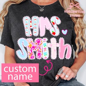 以下が含まれることがあります： 「Mrs. Smith」の文字がパッチワーク風にデザインされた濃いグレーのTシャツ。ピンク、ブルー、ホワイトの要素が含まれており、下部には「custom name」と書かれたピンク色の長方形があります。ハートのグラフィックも表示されています。