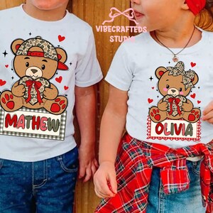 May include: White t-shirts featuring cartoon teddy bears with leopard print accents. One shirt has the name "MATHEW" and the other has "OLIVIA" on a sign. Red hearts and a plaid bow complete the design.