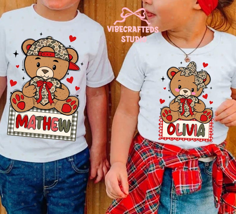 May include: White t-shirts featuring cartoon teddy bears with leopard print accents. One shirt has the name "MATHEW" and the other has "OLIVIA" on a sign. Red hearts and a plaid bow complete the design.