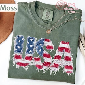 Può includere: Una maglietta verde muschio con la scritta "USA" applicata sul davanti con un design a bandiera americana invecchiata. La maglietta ha un girocollo e maniche corte. Una collana d'argento con un ciondolo è drappeggiata sulla maglietta.