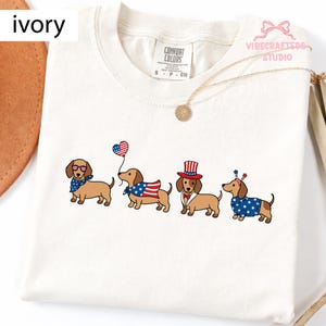 Puede incluir: Camiseta de color marfil con cuatro perros salchicha con atuendos patrióticos. Uno lleva gafas de sol y un pañuelo, otro una bandera, un tercero un sombrero de copa y el último un suéter con estrellas. La camiseta tiene una etiqueta Comfort Colors.