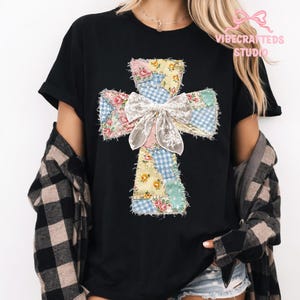 May include: Black t-shirt featuring a patchwork cross design. The cross is made of floral and gingham fabric patches in pastel colors, with a large white bow at the center. The shirt has short sleeves.