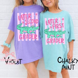 Könnte beinhalten: Zwei Kinder tragen übergroße T-Shirts. Eines ist violett, das andere ist kreidige Minze. Jedes Shirt hat ein rosa-weiß gestreiftes Design mit dem Text "Later Second Grader" und "Later First Grader" mit einem Cartoon-Alligator.