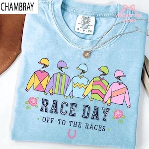 May include: A light blue t-shirt featuring a graphic of five jockey uniforms in various colors and patterns, with the text "RACE DAY" and "OFF TO THE RACES" printed in dark blue, accented by two pink roses and a small pink horseshoe icon.