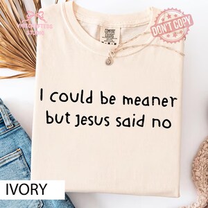 May include: Ivory-colored t-shirt with the text "I could be meaner but Jesus said no" in black. The shirt has a crew neck and a small pendant necklace. The words "Comfort Colors" are printed on the tag.