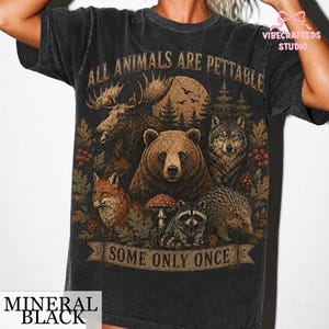 Pu&ograve; includere: Una t-shirt a maniche corte color nero minerale con una stampa grafica raffigurante un alce, un orso, un lupo, una volpe, un procione e un riccio in un bosco. Il testo ALL ANIMALS ARE PETTABLE &egrave; in alto e SOME ONLY ONCE &egrave; in basso.