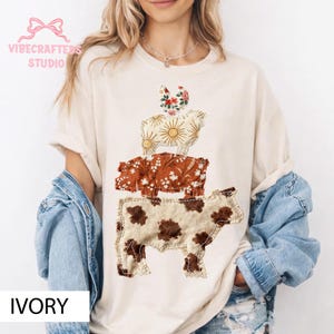 Puede incluir: Camiseta de color marfil con un diseño de animales apilados. El diseño incluye una vaca, un cerdo y una oveja, cada uno con patrones y texturas únicos. La vaca tiene manchas marrones, el cerdo tiene un estampado floral y la oveja tiene un diseño de rayos de sol. La camiseta tiene el texto "IVORY".