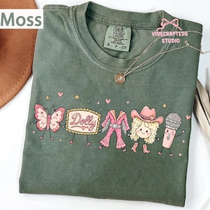 May include: A moss green t-shirt featuring a graphic print of a butterfly, a marquee sign with the text Dolly, a pink fringe outfit, a blonde character wearing a cowboy hat, and a microphone, all holding hands with small stars and hearts.