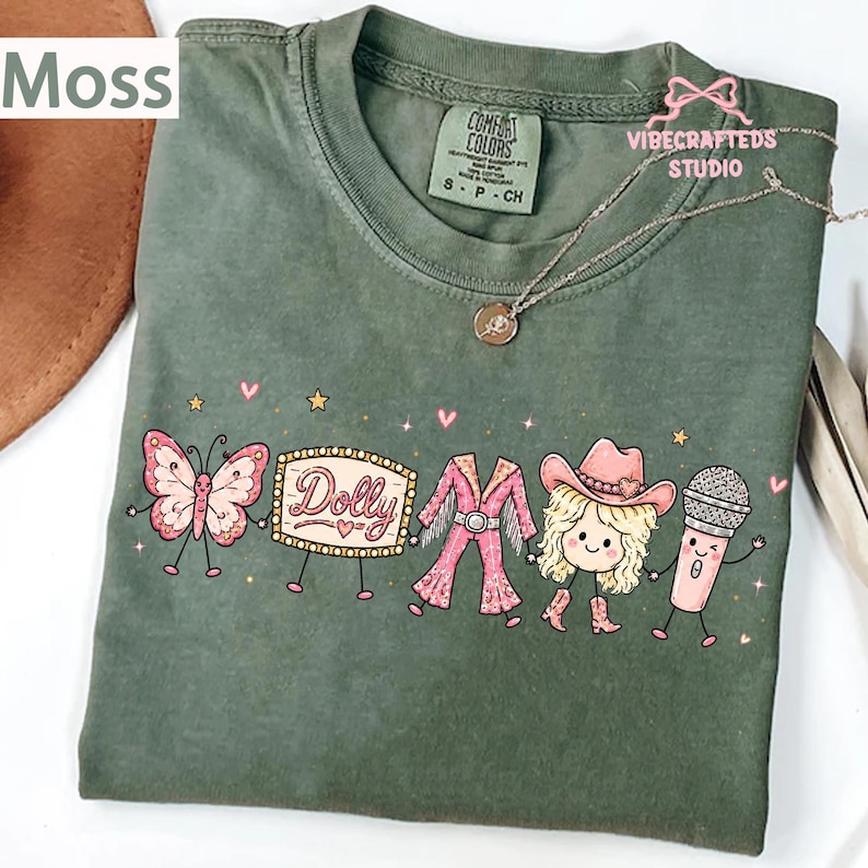 May include: A moss green t-shirt featuring a graphic print of a butterfly, a marquee sign with the text Dolly, a pink fringe outfit, a blonde character wearing a cowboy hat, and a microphone, all holding hands with small stars and hearts.