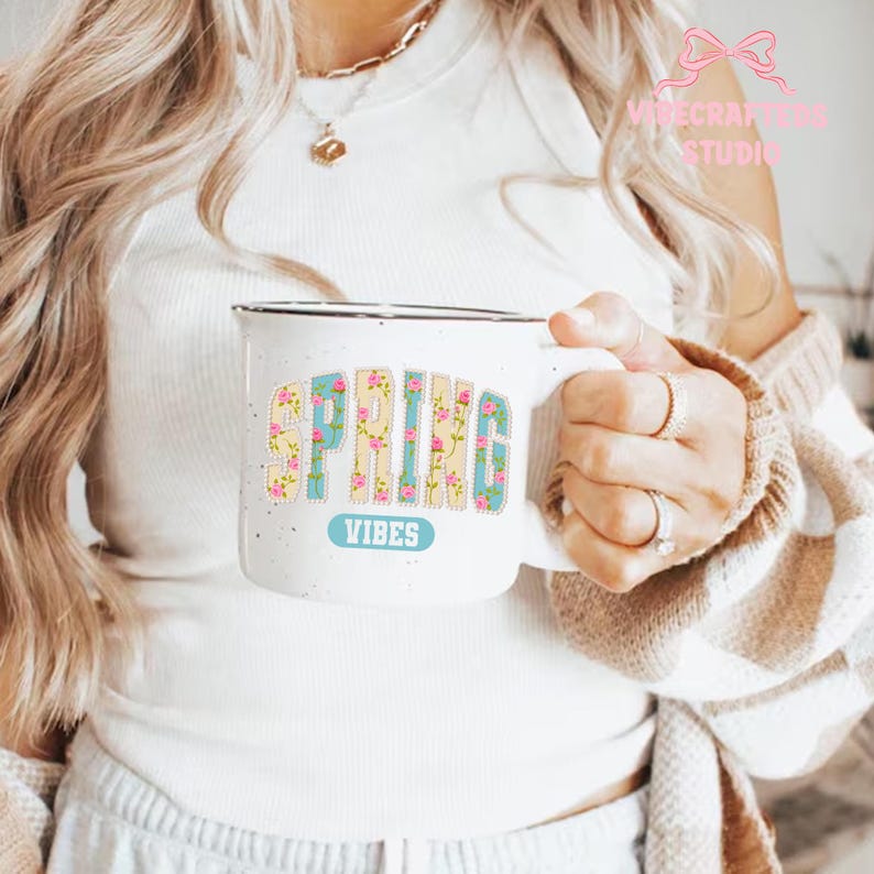 May include: White ceramic mug with the words "SPRING VIBES" in a floral design. The word "SPRING" is in a yellow and pink floral pattern, and "VIBES" is in a light blue color. The mug has a black rim.