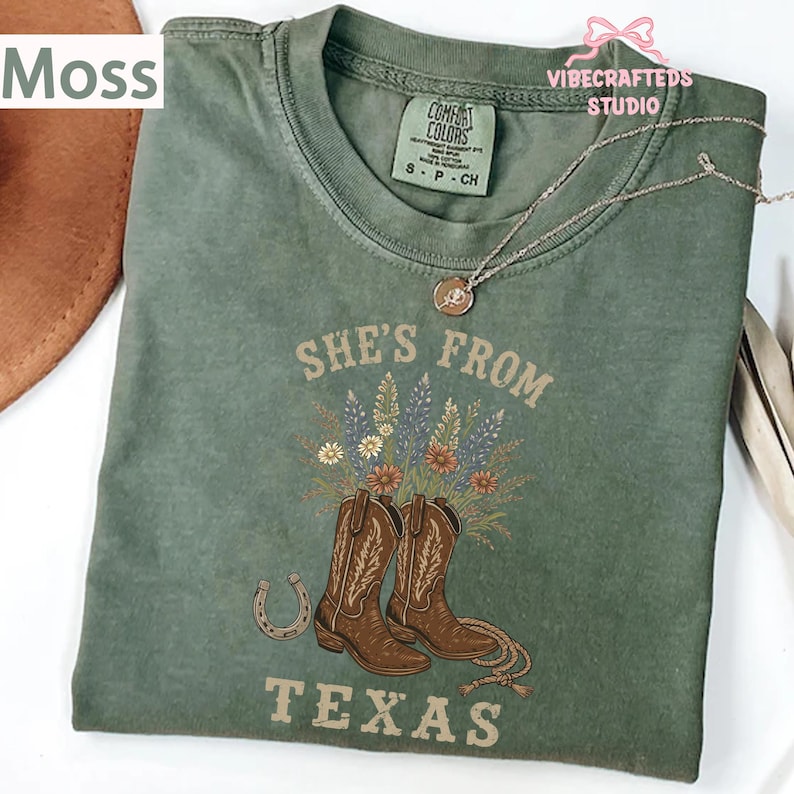 May include: Moss green t-shirt with the text "She's From Texas" and a graphic of cowboy boots and flowers. The shirt has a crew neck and short sleeves. A silver necklace with a pendant is draped across the shirt.