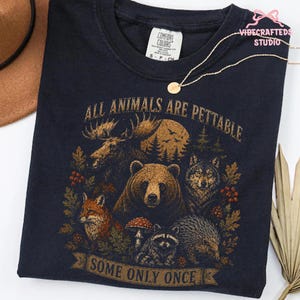 Pu&ograve; includere: Una t-shirt blu navy scuro con una stampa grafica raffigurante un alce, un orso, un lupo, una volpe, un procione e un riccio circondati da elementi boschivi. Il testo recita ALL ANIMALS ARE PETTABLE SOME ONLY ONCE. Etichetta Comfort Colors.