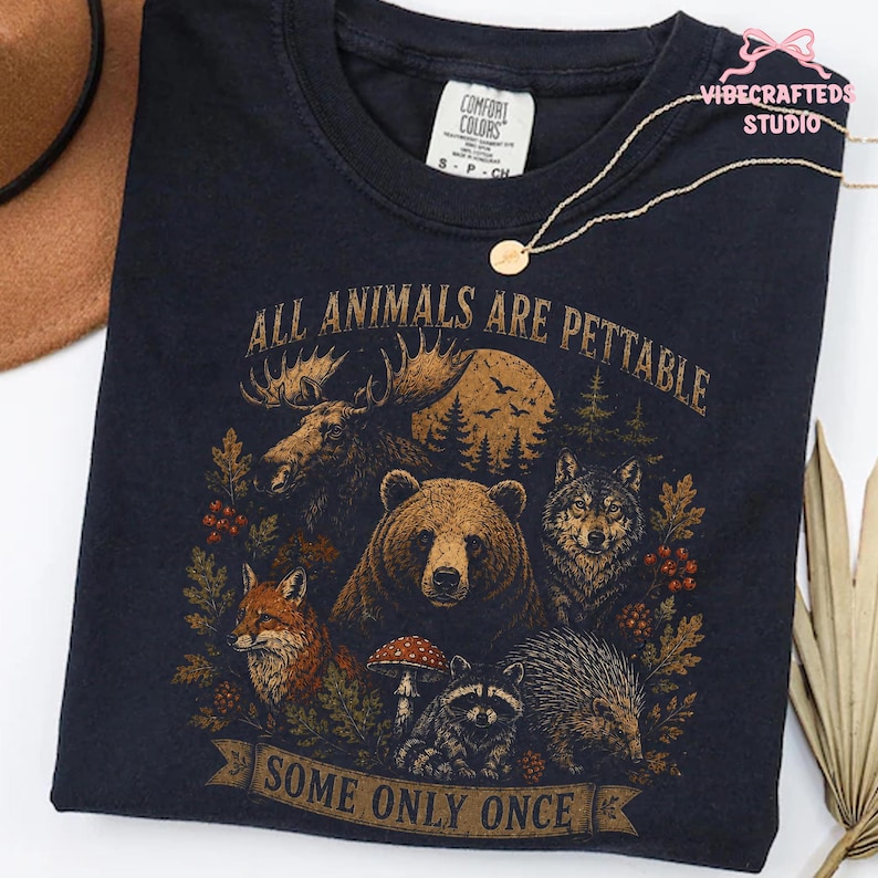 Pu&ograve; includere: Una t-shirt blu navy scuro con una stampa grafica raffigurante un alce, un orso, un lupo, una volpe, un procione e un riccio circondati da elementi boschivi. Il testo recita ALL ANIMALS ARE PETTABLE SOME ONLY ONCE. Etichetta Comfort Colors.