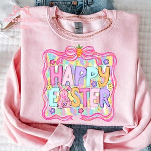 May include: A light pink sweatshirt with a colorful "Happy Easter" graphic. The graphic features a bunny, flowers, and a bow. The text is in pastel colors, with a carrot on top. A gold chain is around the neck.