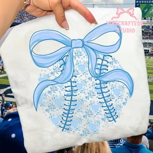 May include: A white baseball-shaped design with a light blue bow and stitching. The baseball has a floral pattern in light blue. The text "Wire Crafted Studio" is visible in the upper right corner.