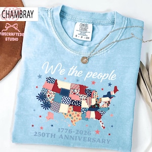 May include: Pale blue t-shirt featuring the text "We the people" and a patchwork map of the United States. The map is decorated with red, white, and blue patterns. The shirt also features the text "1776-2026 250th Anniversary".