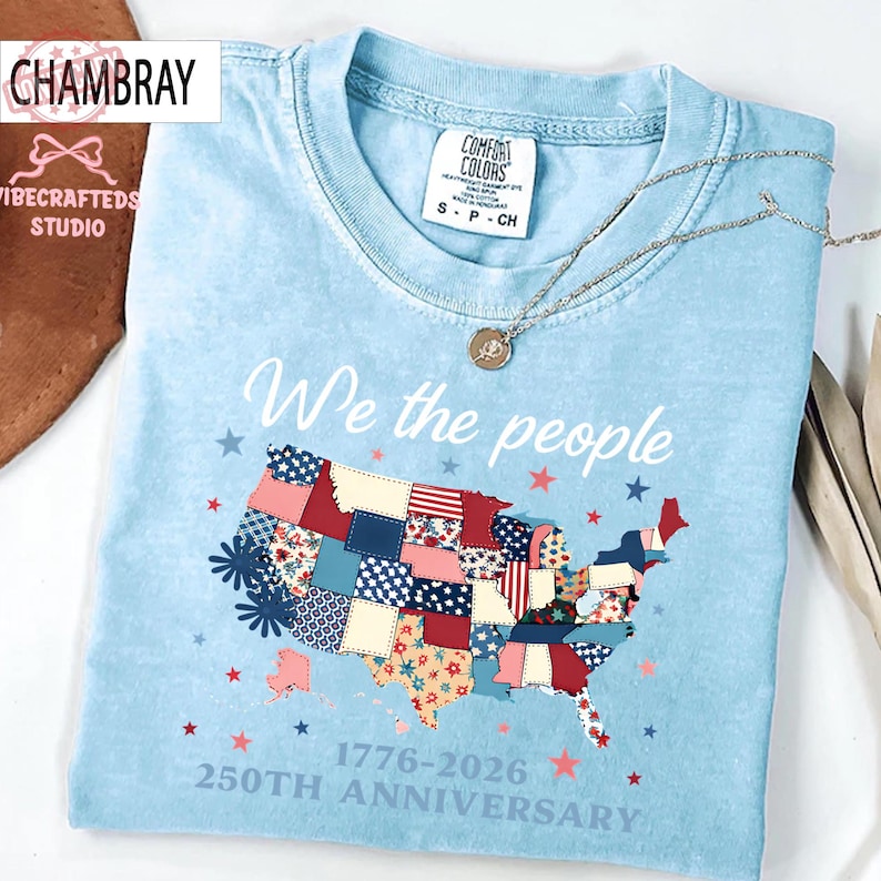 May include: Pale blue t-shirt featuring the text "We the people" and a patchwork map of the United States. The map is decorated with red, white, and blue patterns. The shirt also features the text "1776-2026 250th Anniversary".
